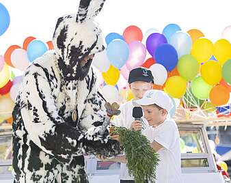 Mann in Hasenkostüm übernimmt von zwei Kindern ein Bund Karotten im Hintergrund Luftballons
