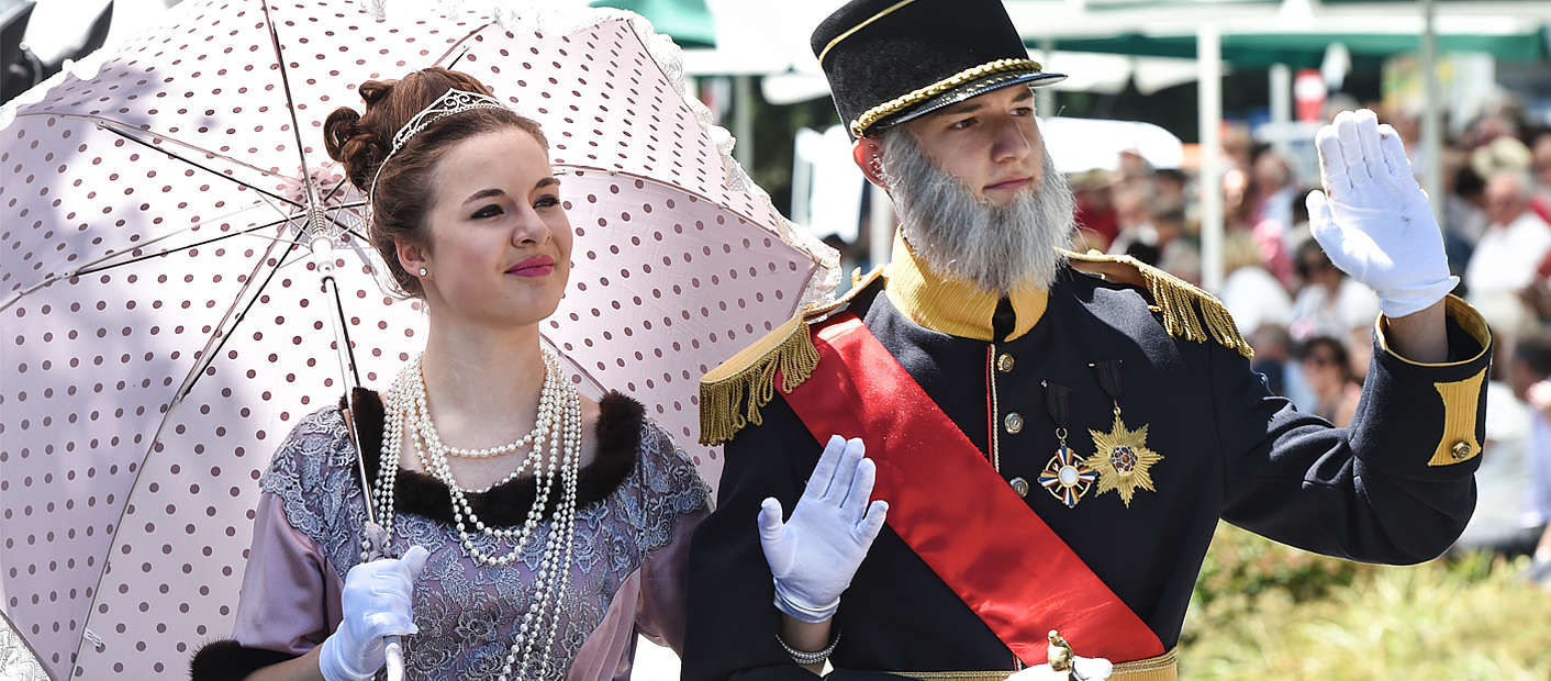 junge Frau verkleidet als Dame mit Sonnenschirm und Perlenkette und junger Mann verkleidet als Mann in Uniform mit Scherpe und Abzeichen
