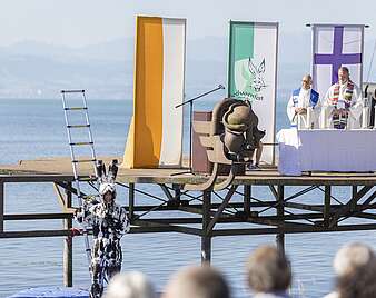 Menschen verfolgen ökumenischen Freiluftgottesdienst mit Altar auf Klangschiff, Mann in Hasenkostüm winkt