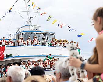 Seehas winkend auf einem Schiff, links und rechts von ihm Fanfarenzug