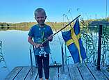 Junge hält Seehasen-Kuscheltier und schwedische Nationalflagge in den Händen und steht vor See.