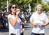 Festzug: Frau und Mann moderieren auf Straße, Frau spricht in Mikrofon